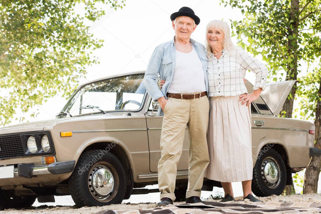 bottom view of senior couple standing near beige vintage car and looking at camera 