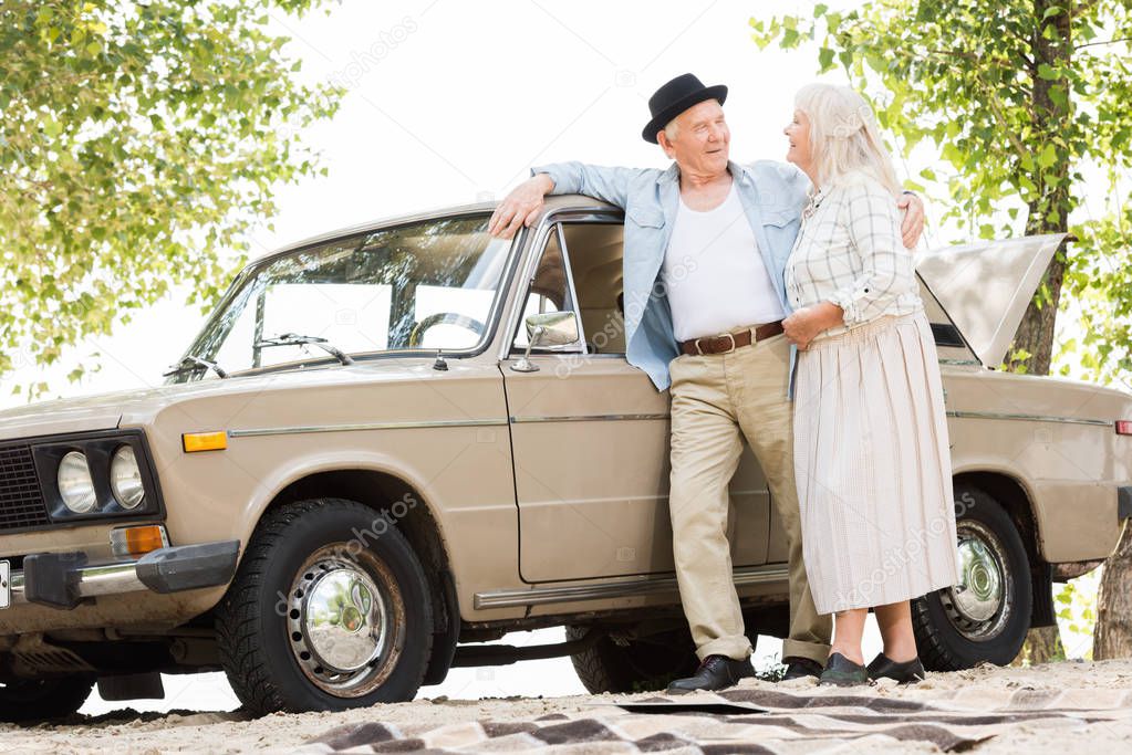 bottom view of stylish senior couple standing near beige vintage car and looking at each other 