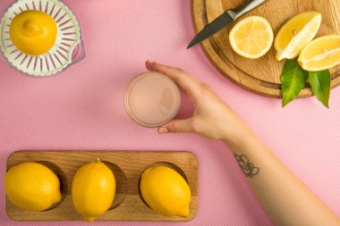 top view of female hand with glass of lemon juice on pink table with squeezer and lemons on wooden boards clipart