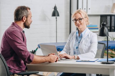 smiling female physiotherapist typing on laptop during appointment with patient in hospital clipart