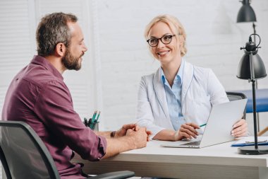 smiling chiropractor pointing at laptop screen during appointment with patient in hospital clipart