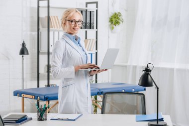 gözlük ve beyaz önlük chiropractor Kliniği içinde laptop ile gülümseyen yan görünümü