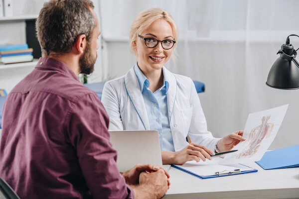 улыбающаяся женщина-хиропрактик показывает пациенту фотографию человеческого тела во время приема в больнице

