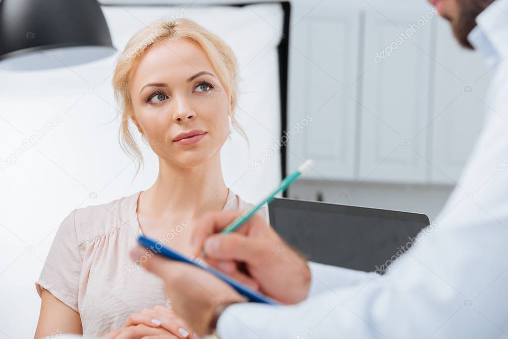partial view of physiotherapist in white coat making notes in notepad with female patient near by during appointment in clinic