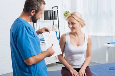 physiotherapist showing human body scheme to woman on massage table in hospital clipart