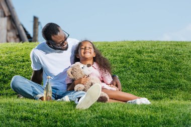 Afro-Amerikan baba ve soda ve oyuncak ayı eğlence parkı ile Hill'de oturan gülümseyen kız