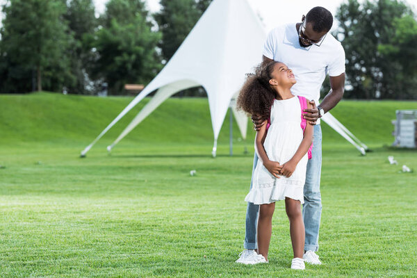 african american father hugging daughter with school bag in park