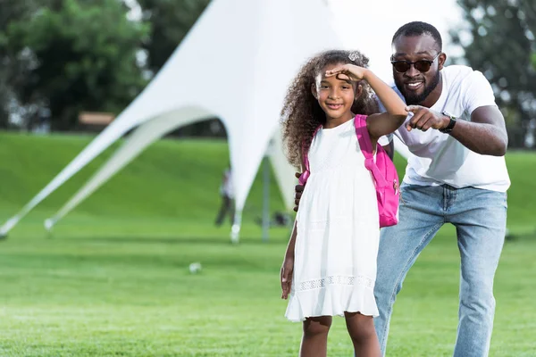 非洲裔美国人的父亲指向照相机对女儿与书包在公园 — 图库照片