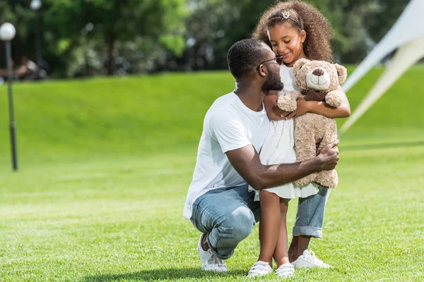 Afroamericano Padre Accovacciato Abbracciare Adorabile Figlia Con Orsacchiotto Nel Parco — Foto Stock