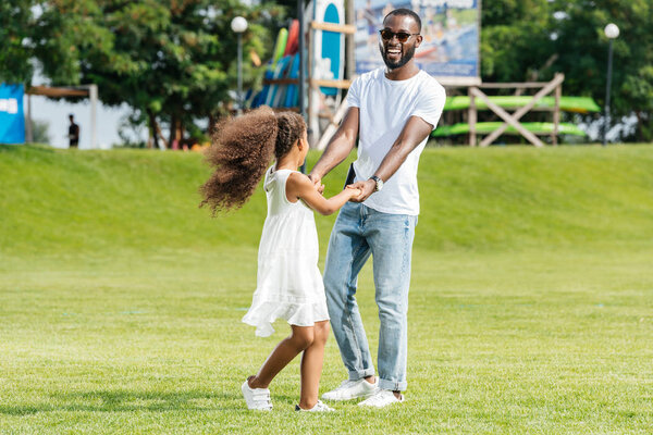 happy african american father and daughter holding hands and spinning around in amusement park