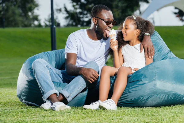非洲裔美国人父亲舔冰淇淋和坐在豆子椅与女儿在公园 — 图库照片
