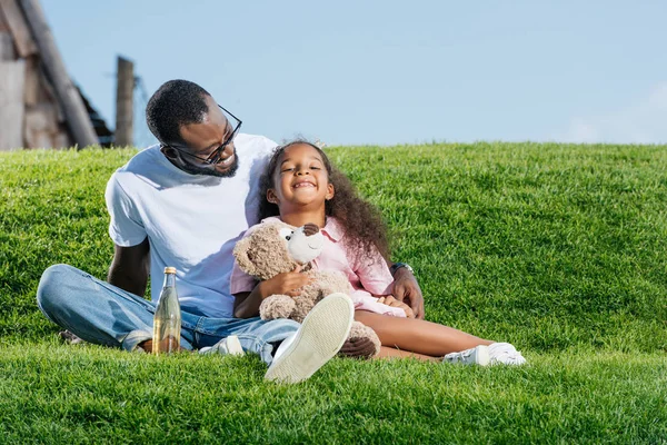 アフリカ系アメリカ人の父とソーダと遊園地でテディベアの丘の上に座って笑顔の娘 — ストック写真