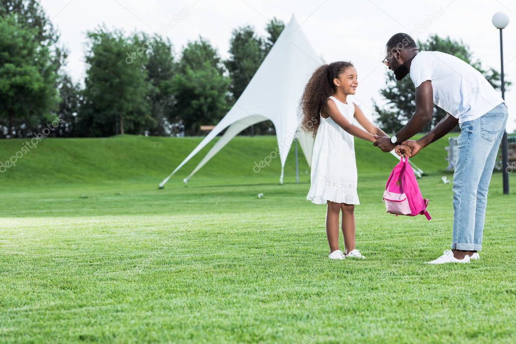 african american daughter giving school bag to father in park