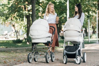 mothers holding coffee to go and walking with baby strollers in park clipart