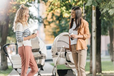 mothers talking, standing with coffee to go and baby strollers in park  clipart