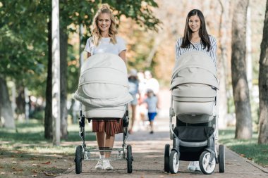 smiling mothers walking with baby strollers in park and looking at camera clipart