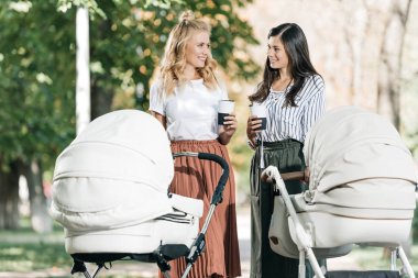 mothers holding disposable coffee cups and looking at each other near baby strollers in park clipart
