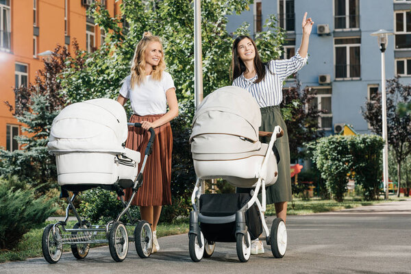 mothers walking with baby strollers on street, woman pointing on something to friend