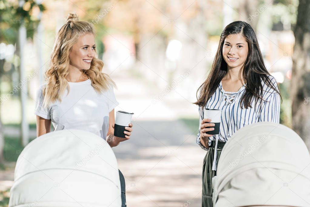 smiling mothers standing with baby strollers and coffee in paper cups in park