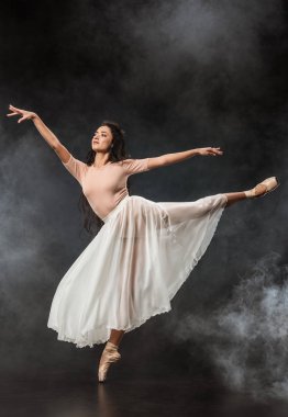 beautiful young ballerina in white skirt dancing on dark background with smoke around clipart