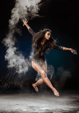 beautiful ballerina in black bodysuit jumping on dark background with talc powder around clipart