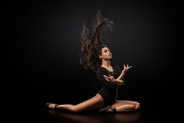 young attractive ballerina in black bodysuit and ballet shoes posing on dark backdrop