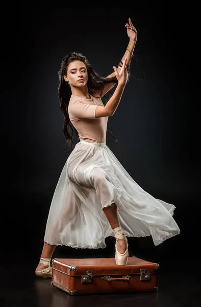 美丽的芭蕾舞演员在白色裙子站立与一条腿在复古手提箱在黑暗的背景上 — 图库照片