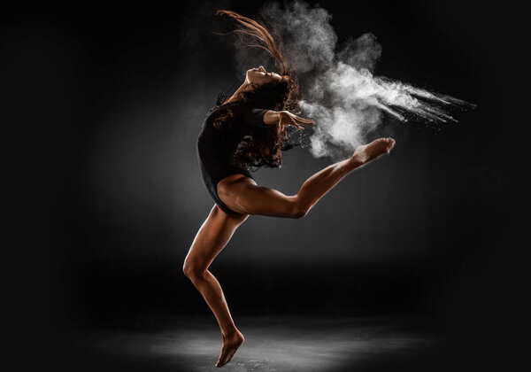 молодая балерина в черном купальнике с тальковым порошком танцует на темном фоне
