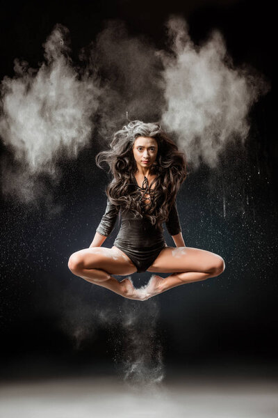 beautiful ballerina in black bodysuit jumping on dark background with talc powder around