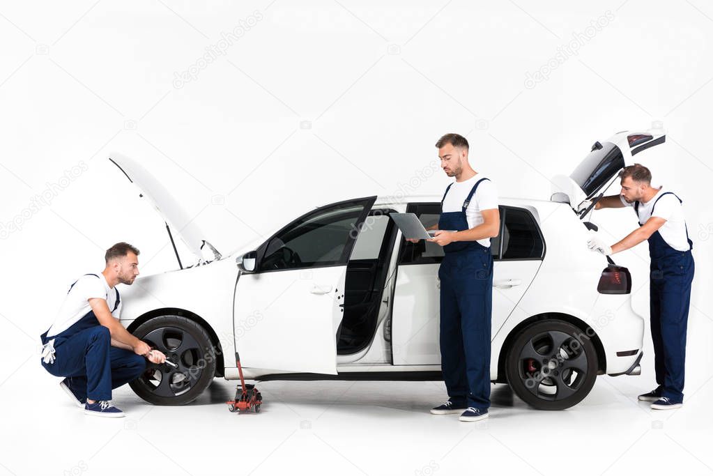 auto mechanic changing car tire, using laptop and looking in open car trunk on white