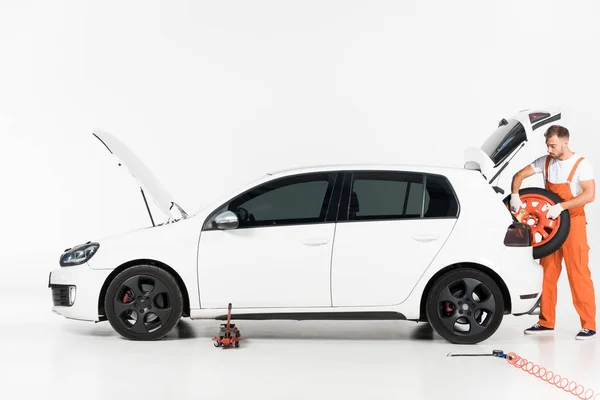 auto mechanic in orange uniform taking tire from car trunk on white