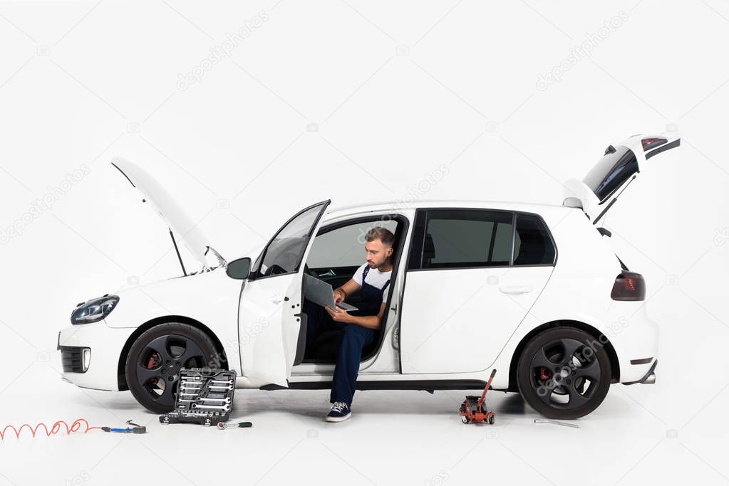 handsome auto mechanic sitting in broken car and using laptop on white