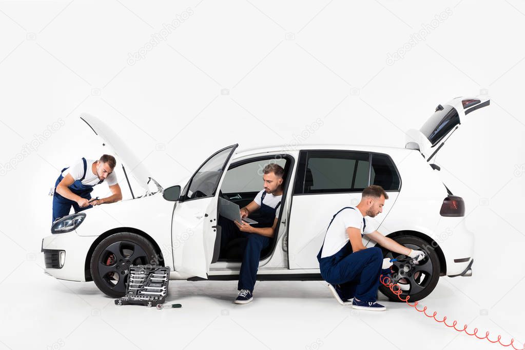handsome auto mechanic inflating tire, using laptop and looking in hood on white