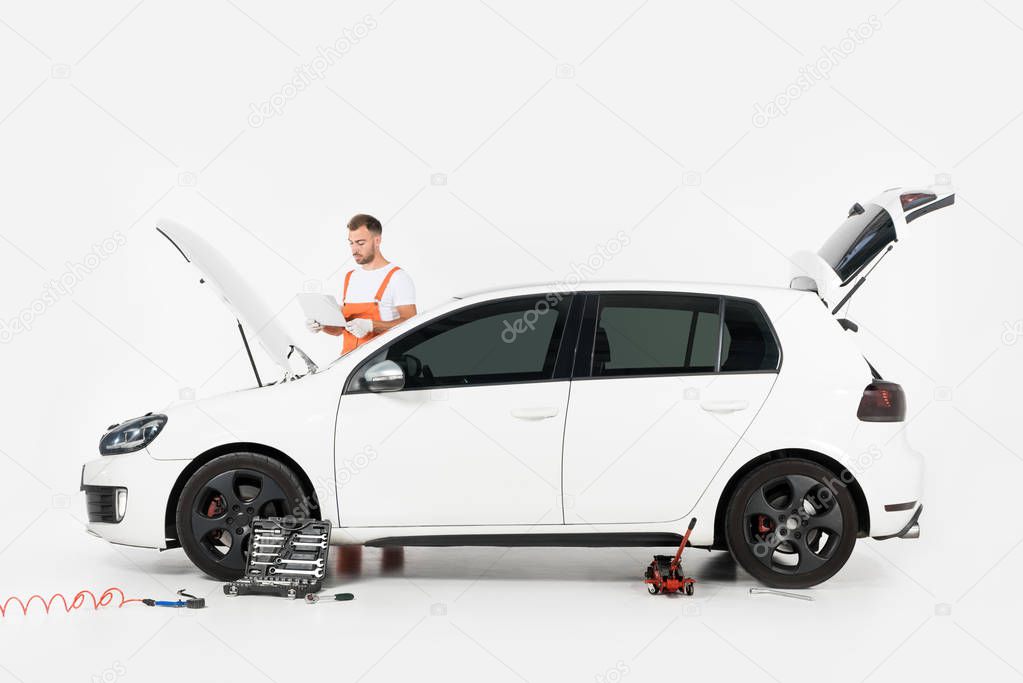 handsome auto mechanic using laptop near car with open hood on white