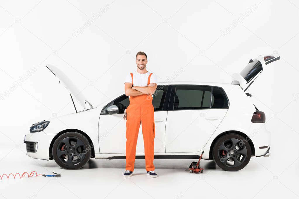 smiling handsome auto mechanic standing with crossed arms near broken car on white