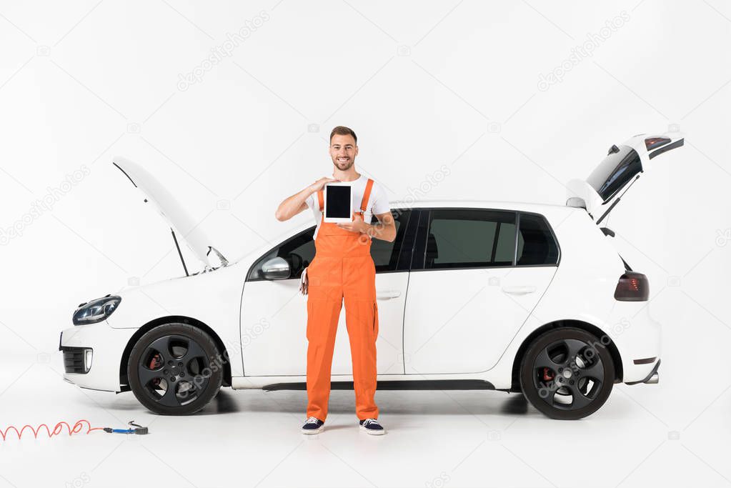 handsome auto mechanic showing tablet with blank screen near broken car on white