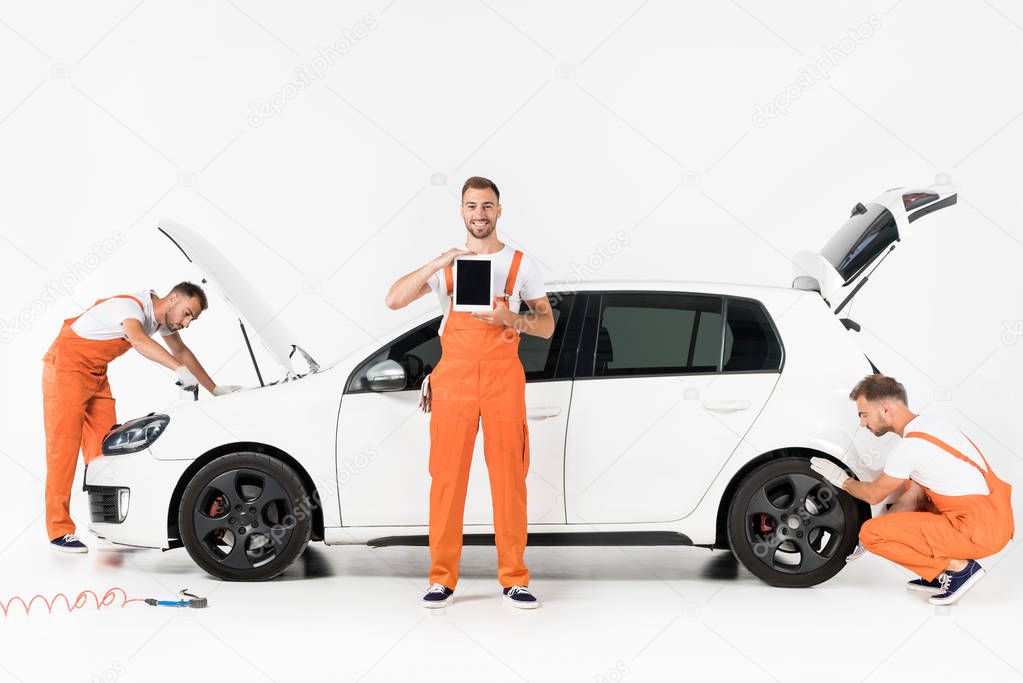 auto mechanic changing car tire, showing tablet and looking in open car hood on white