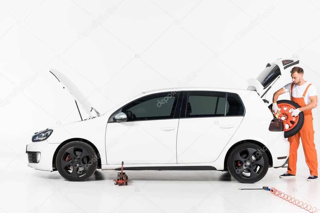 auto mechanic in orange uniform taking tire from car trunk on white