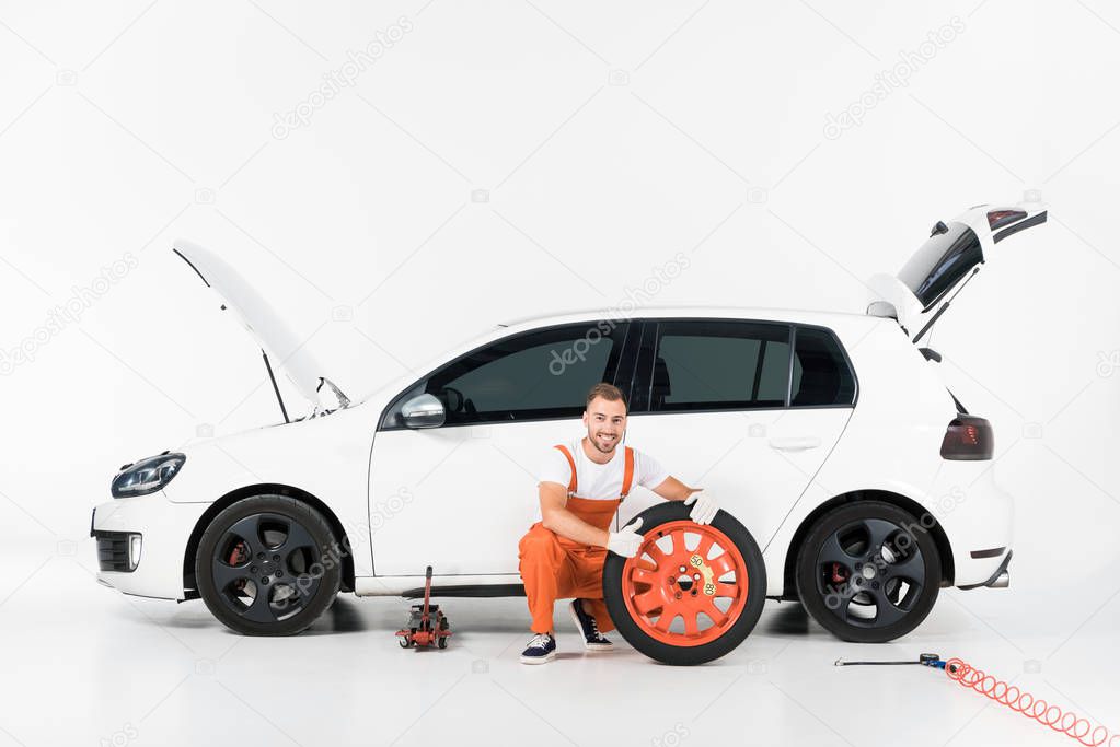 auto mechanic squatting near car tire and showing thumb up on white