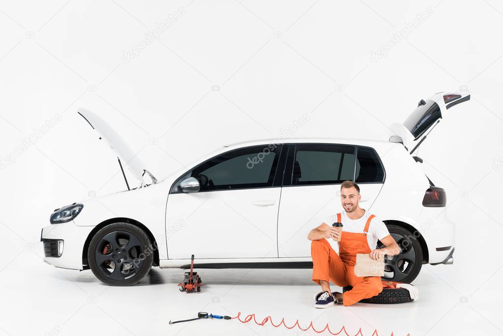handsome auto mechanic sitting near car with coffee in paper cup and lunch in paper bag on white