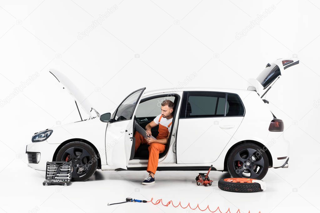 handsome auto mechanic using laptop in broken car on white
