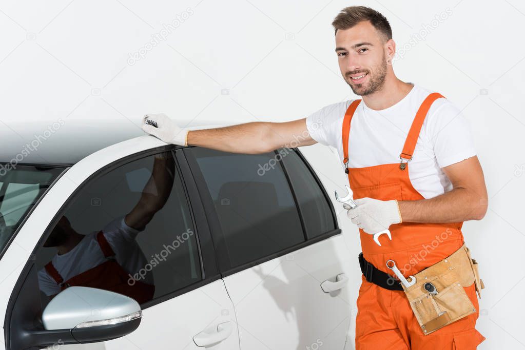 handsome auto mechanic holding wrench and leaning on car on white