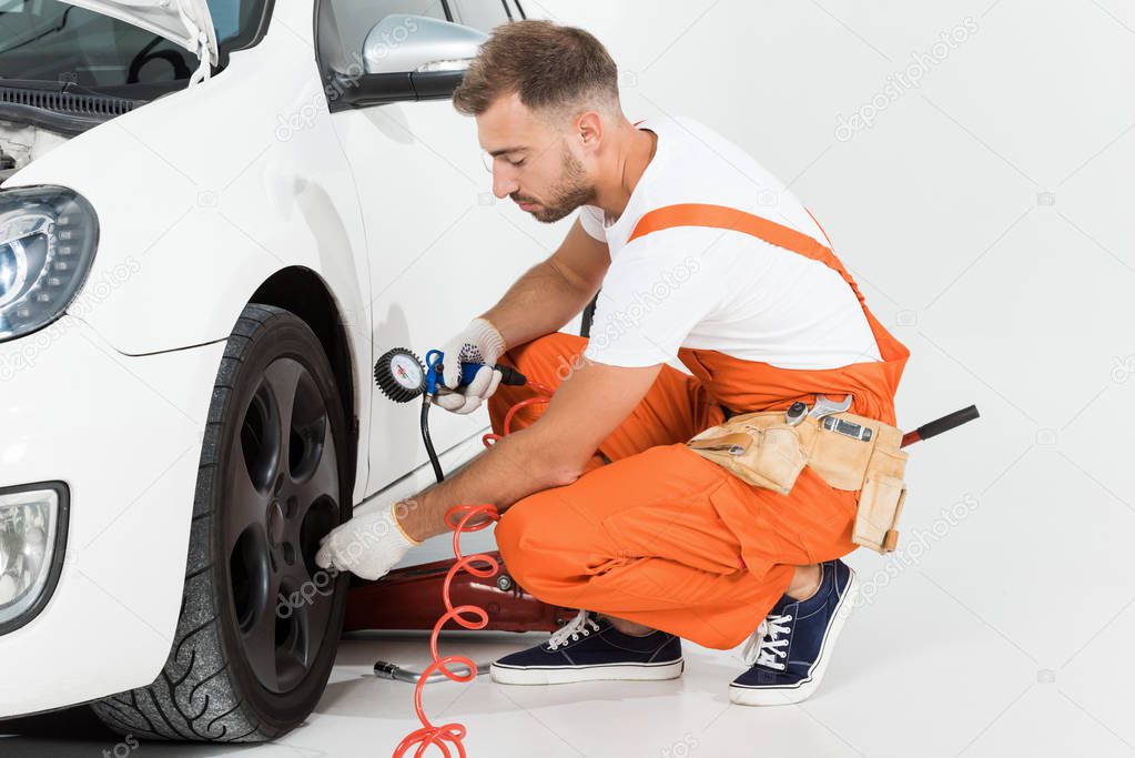 handsome auto mechanic inflating tire and checking air with gauge pressure on white