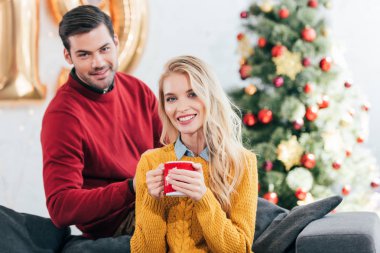 kahve fincanı yakınındaki evde ile Noel ağacı oturan adam tutan gülümseyen kadın
