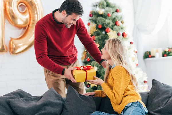 Stilig Man Gifting Närvarande Till Kvinna Hemma Med Julgran — Gratis stockfoto