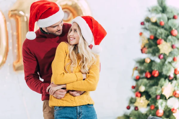 Hermosa Pareja Sombreros Santa Abrazándose Mirándose Casa Con Árbol Navidad —  Fotos de Stock