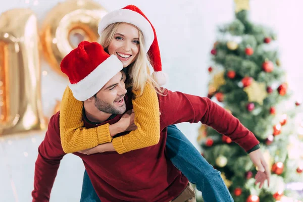 Happy Man Giving Piggyback His Smiling Girlfriend Home Christmas Tree — Stock Photo, Image