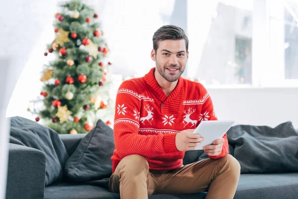 デジタル タブレットを使用して 背後にあるクリスマス ツリーが付いているソファーの上に座って幸せな男 — ストック写真