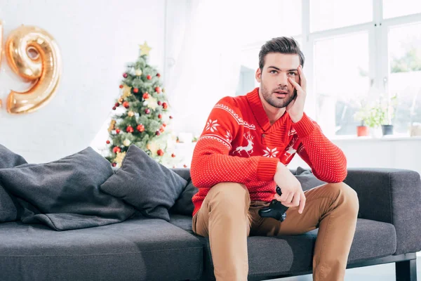 upset man holding joystick and sitting on sofa with christmas tree behind