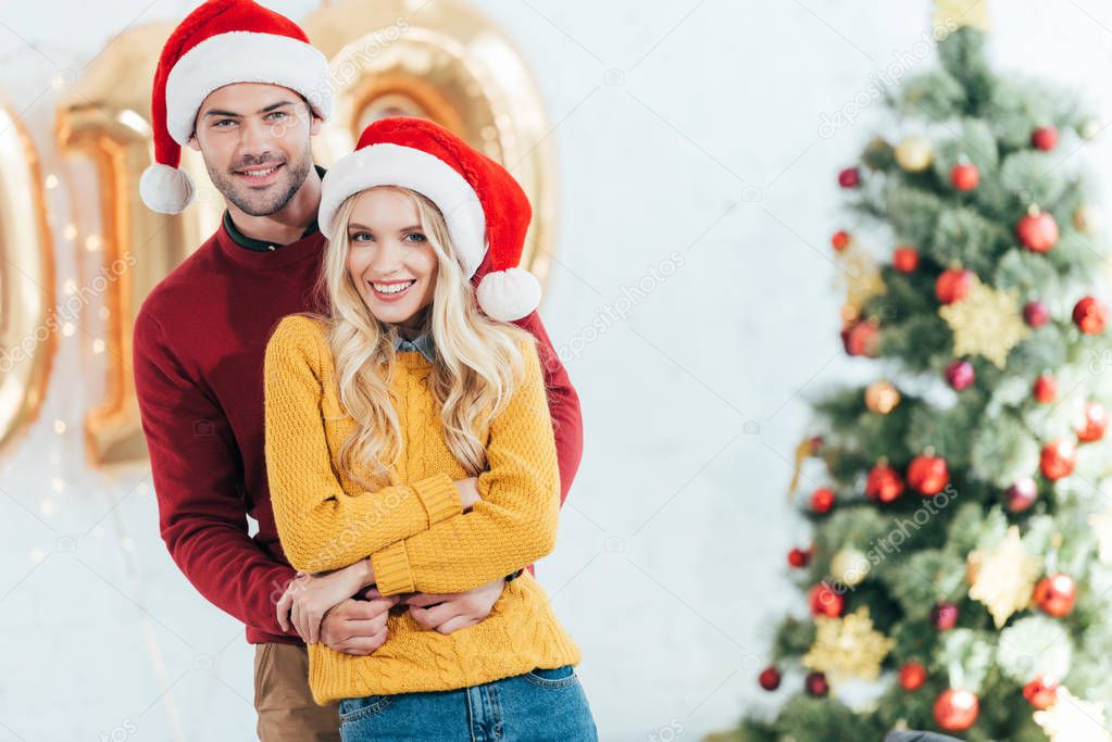 happy couple in santa hats embracing at home with christmas tree